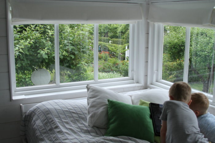 Two children are lying in bed reading, with a green garden visible through the windows.