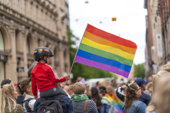 A young boy sitting on the shoulders of an adult and waving a big rainbow flag amidst other parade-goers.