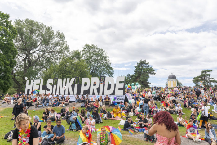 A large crowd of people sitting on the lawn in Kaivopuisto park, a big Helsinki Pride sign in the background.