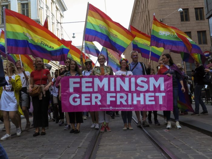 A group of people walking on the street waving rainbow flags and holding a large banner saying 'Feminism gives you freedom' in Swedish.