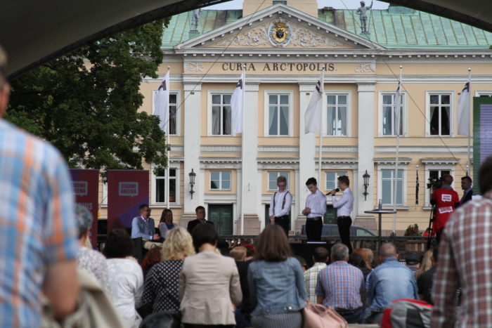 Three young people talking on a stage to a three-person jury with a crowd watching.