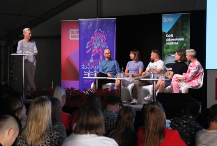 Five people and a host gathered for a panel discussion on a small stage with a small crowd watching.