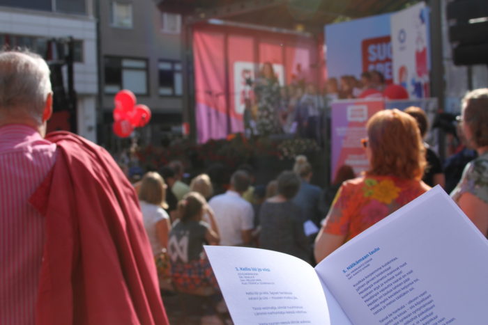 A person holding a leaflet with song lyrics in a crowd gathered by a small stage.