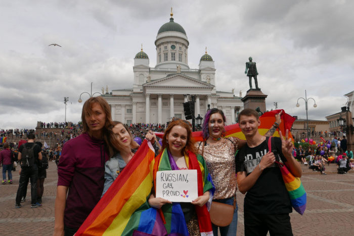 Five people with rainbow flags standing in front of the Helsinki cathedral; the person in the middle is holding a sign saying 'Russian, gay and proud'.