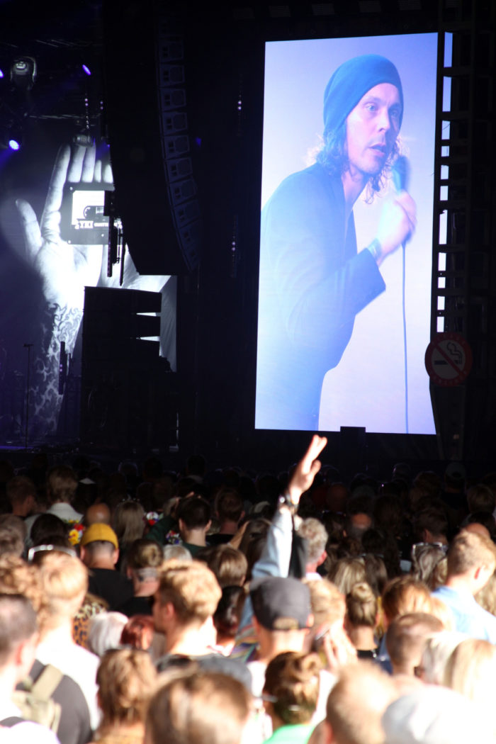 A crowd listens to a Ville Valo concert, with Valo visible on a huge screen by the stage.