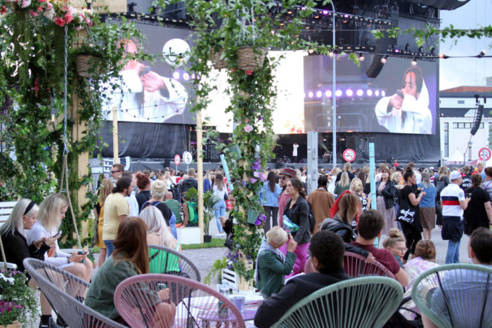 People chill in café chairs while rapper Earl Sweatshirt is visible on screens by a stage in the background.