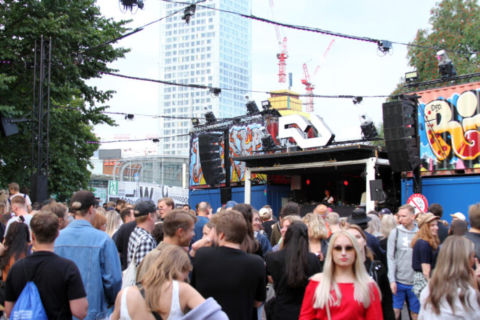 A crowd gathers by a small stage surrounded by freight containers covered in graffiti.