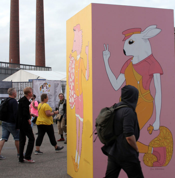 Festival-goers admire artwork, paintings on enormous boxes showing animals wearing clothes.