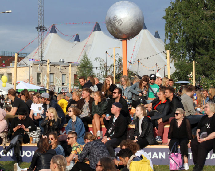 A crowd of festival-goers sits on benches, with an enormous white tent in the background.