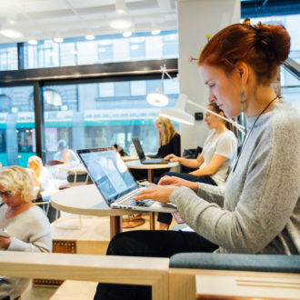 A group of women work on their laptops and phones at a café, with a tram visible through the window.