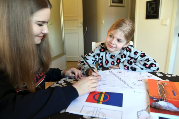 Two girls sit at a table with schoolbooks, notebooks and a colour printout of the Sámi flag. 