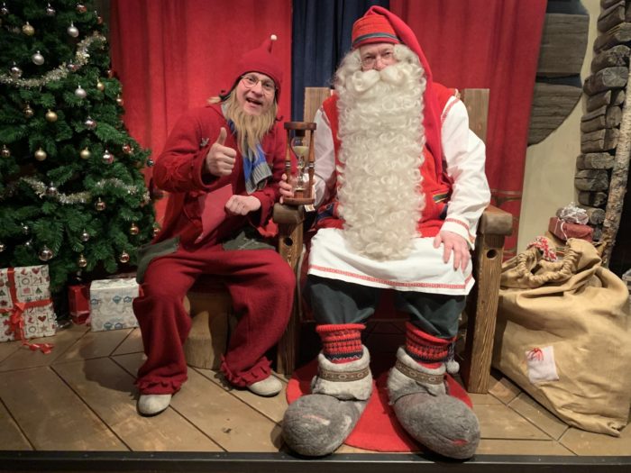 Santa Claus and an elf sit in front of a Christmas tree. The elf is giving a thumbs-up sign.