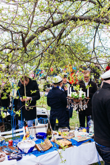 People wearing Finnish high school graduation caps stand near a table stacked with food and drink in a park.