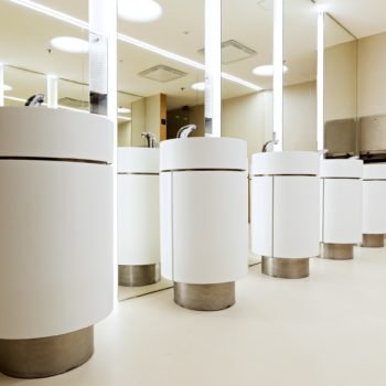 Fünf weiße zylindrische Gebilde mit jeweils einem Waschbecken und einem Wasserhahn stehen in einer Reihe vor Spiegeln in einem Waschraum.