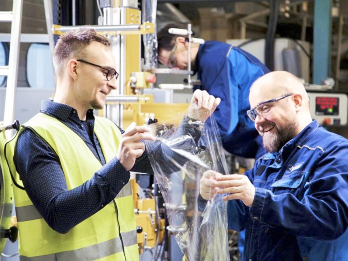 Два мужчины в мастерской, в рабочей одежде и в очках, держат на руках материал, похожий на пластиковую плёнку. 