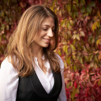 Vor einem herbstlichen Hintergrund aus grünen und roten Blättern lächelt eine Frau in einer weißen Bluse und schwarzen Weste.