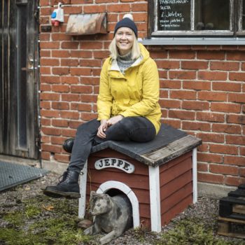 一名女性穿着外衣戴着帽子坐在一座犬舍顶上，犬舍门口有一条狗的塑像。