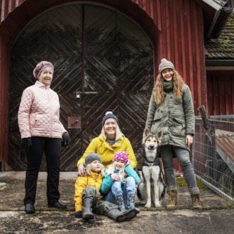 Usando jaquetas e chapéus que sugerem clima frio, uma mulher mais velha, duas mulheres na casa dos trinta e duas crianças pequenas posam juntas com um cachorro grande em frente a um prédio de madeira.