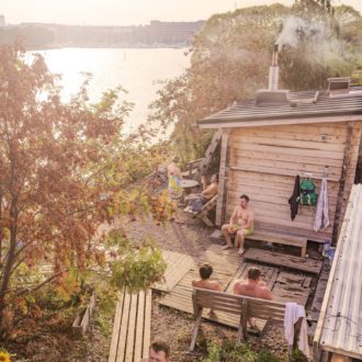Plusieurs personnes à la taille ceinte d’un drap de bain sont assises sur des bancs en bois sur un rivage, tandis qu’on voit à côté une petite maison en bois munie d’une cheminée.
