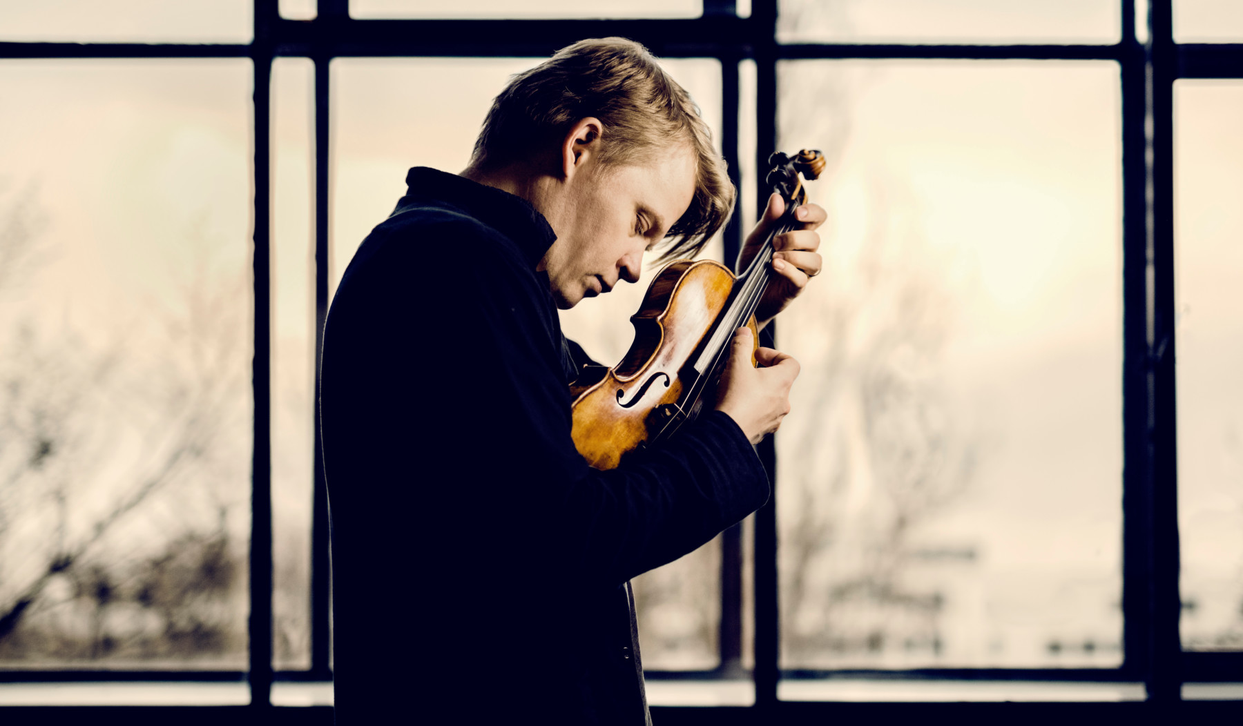 Violinist Pekka Kuusisto strums his violin without a bow in front of a window.