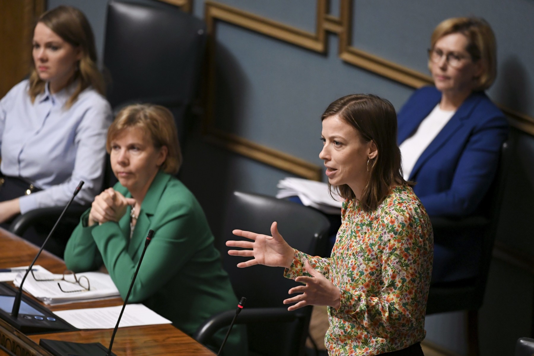 Three women are sitting and listening as a fourth woman delivers a speech before Parliament.