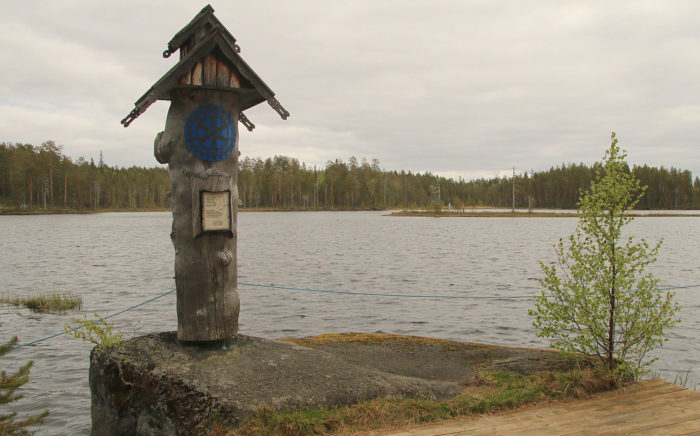 Un poteau en bois se dresse au bord d’un lac au milieu de la campagne. 