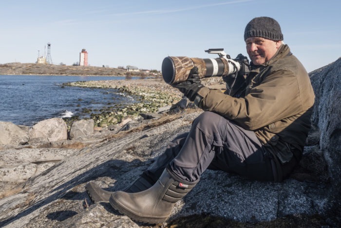 Мужчина сидит у каменистой береговой линии, настраивая фотоаппарат с телеобъективом.