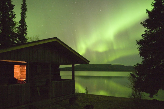 Домик у ночного озера с небом, полным волнистых зеленых узоров северного сияния.