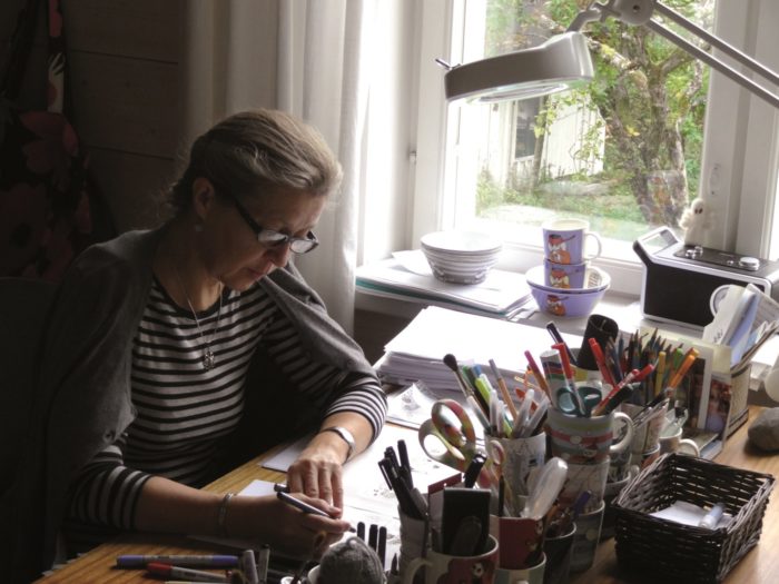 Designer Tove Slotte sits by a desk next to a window, and is drawing on a piece of paper. On the desk, there are Moomin cups filled with scissors and pencils. 