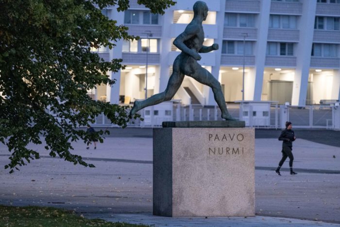 Uma estátua de um homem correndo está em um pedestal em frente a um estádio, e um corredor passa correndo por ela.