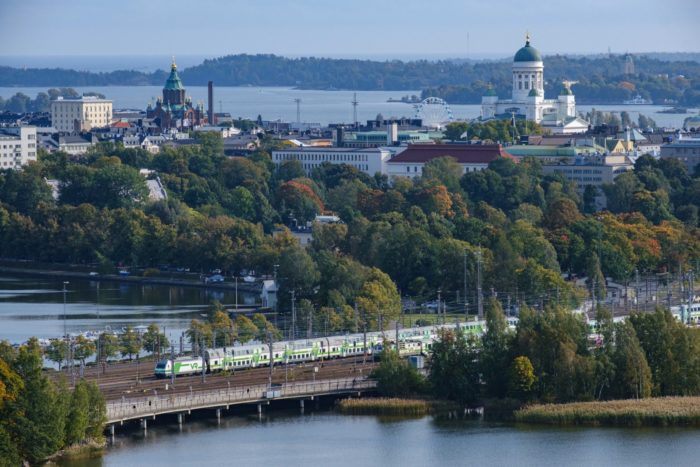 Una vista de Helsinki con la bahía, un tren que pasa, un parque lleno de árboles y cúpulas de iglesias.