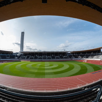 Uma vista dentro de um estádio mostra a pista, o campo e o céu acima da borda do telhado que cobre as arquibancadas.