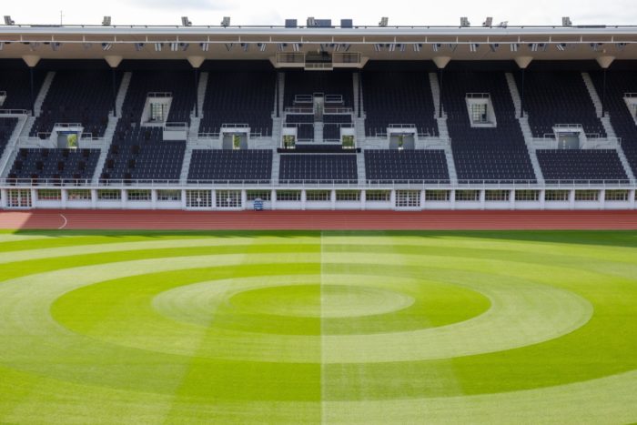 Rows of stadium seating are in the background, behind the grass of a football pitch.