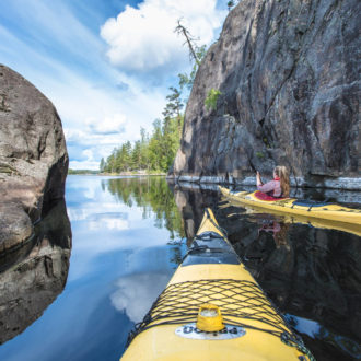 A person in a kayak paddles on a lake with a rocky cliff on one side.