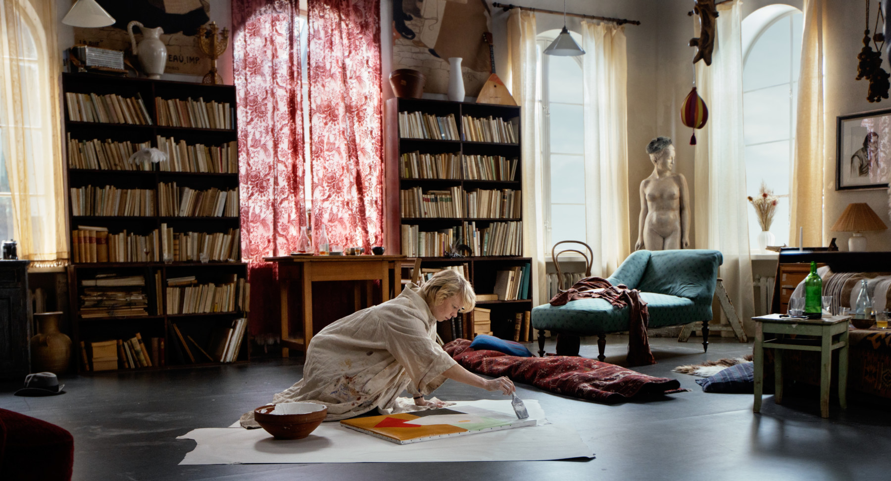 Une femme peint sur une toile étalée sur le plancher d’une vaste pièce où des étagères chargées de livres sont visibles à l’arrière-plan. 
