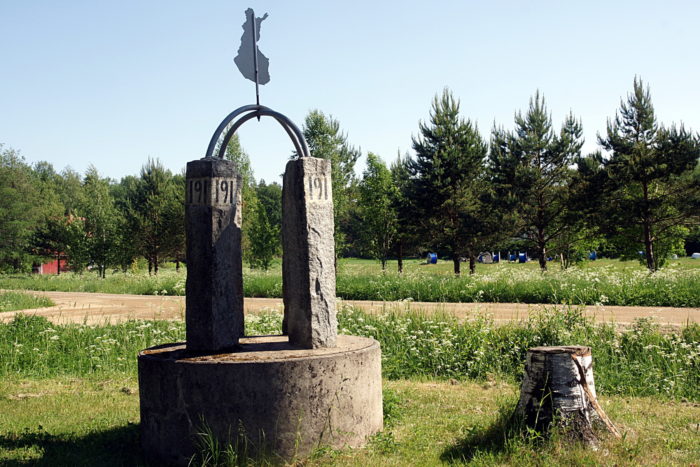 Deux piliers en pierre reliés par des barres métalliques incurvées se dressent à côté d’un tronc d’arbre et d’une petite route de campagne, tandis que des arbres sont visibles à l’arrière-plan. 