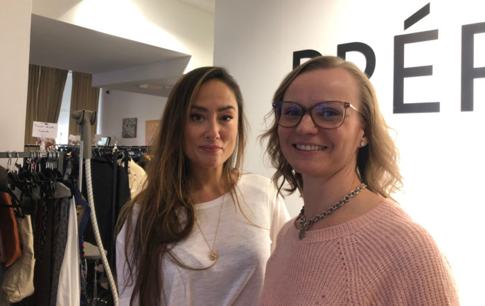 Dans une boutique, deux jeunes femmes prennent la pose pour une photo devant des portants chargés de vêtements. 
