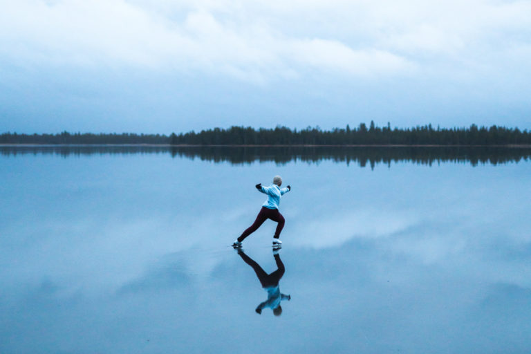A woman ice skates across a lake whose surface reflects the sky.