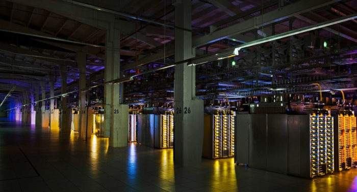 Rows of stacked computer equipment show yellow, blue and orange lights in a dimly lit industrial hall.