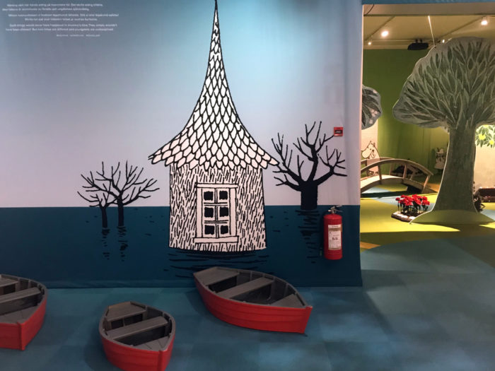 Three small boats are on the floor in front of a picture of a house in deep floodwater.