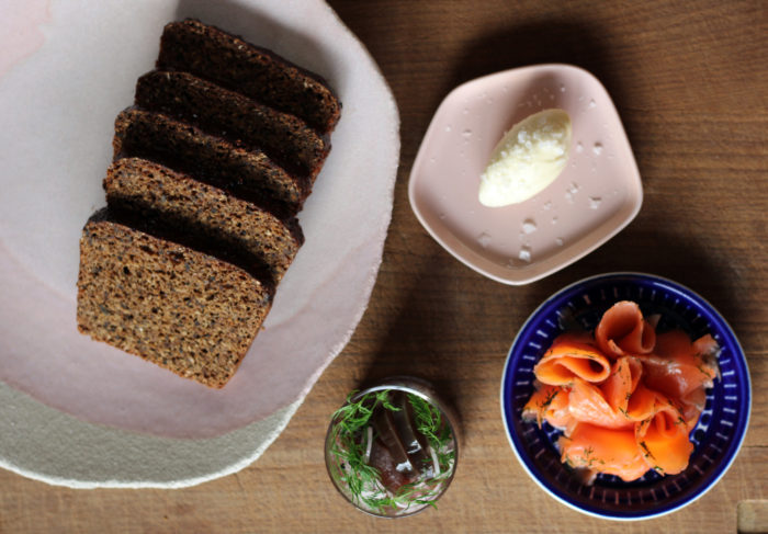 Une grande assiette de pain tranché, une soucoupe où a été déposée une noisette de beurre, une petite assiette garnies de tranchettes de saumon et une verrine pleine de bouchées d’aubergines sont disposées sur une table. 