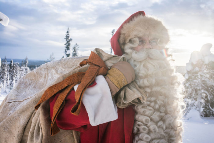 Santa Claus carries a bag over his shoulder, with a snowy forest in the background.