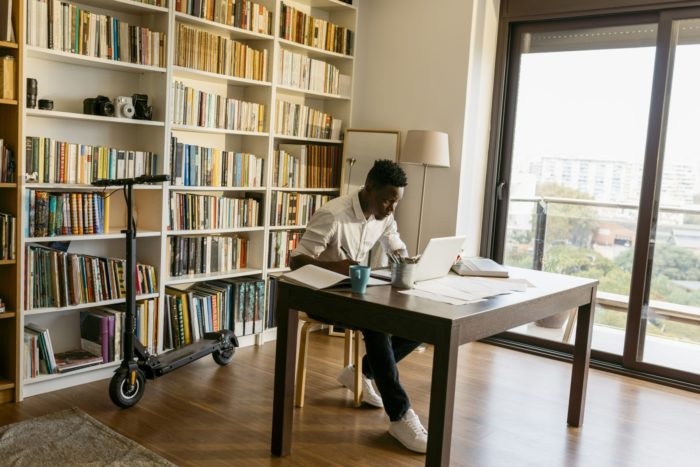 En un apartamento, un hombre está sentado ante un escritorio con un ordenador portátil, libros y papeles.