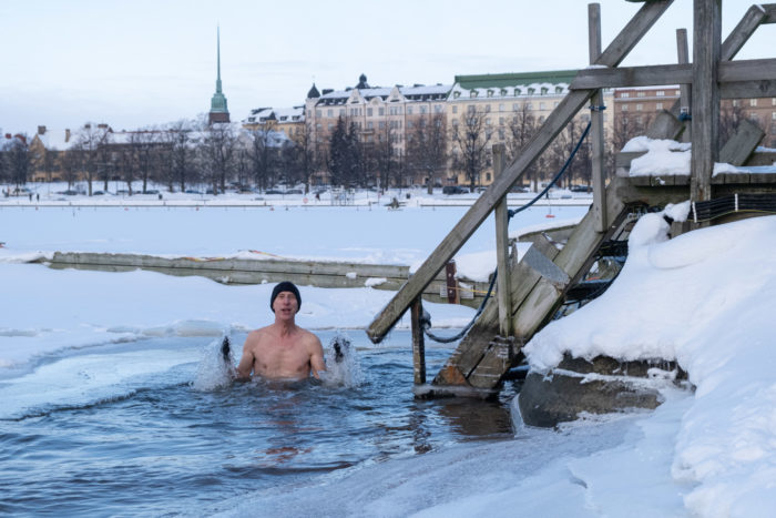 Um homem está em grande buraco no gelo, com água até o peito, rodeado pela neve, com uma escada de saída nas proximidades e uma vista de Helsinque ao fundo.