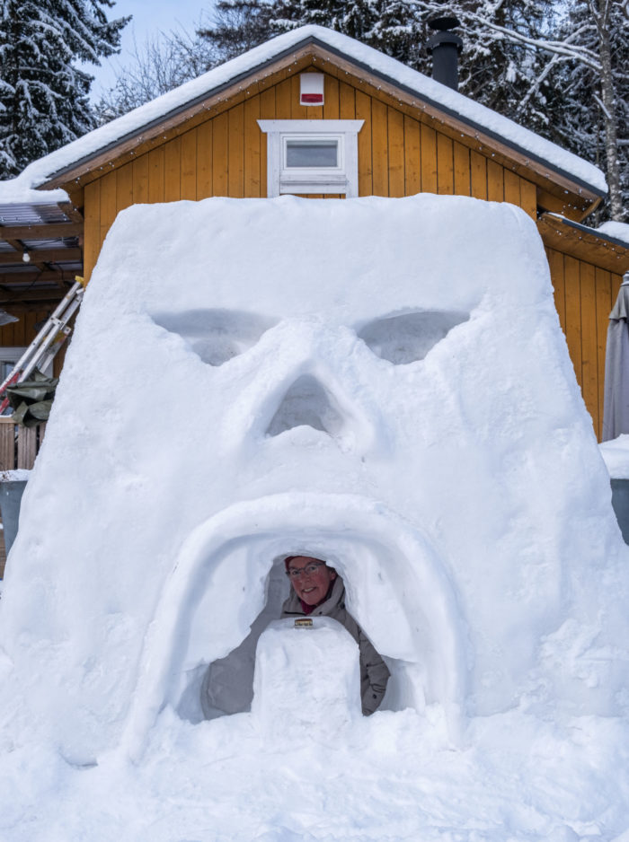 Une personne passe la tête à travers une ouverture creusée au milieu de la façade d’un château de neige dont l’aspect fait plus ou moins penser à une grande tête d’homme affichant une expression mécontente.