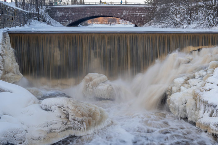 Debout sur un pont, des gens contemplent des formations de glace visibles en aval de l’ouvrage.