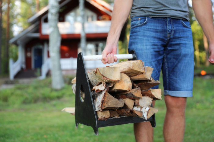Une personne traverse une cour de maison en été en portant un chargement de bûches de bois fraîchement coupées.