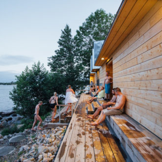 Several people sit, wrapped in towels, on a wooden bench outside a sauna on a rocky shore.