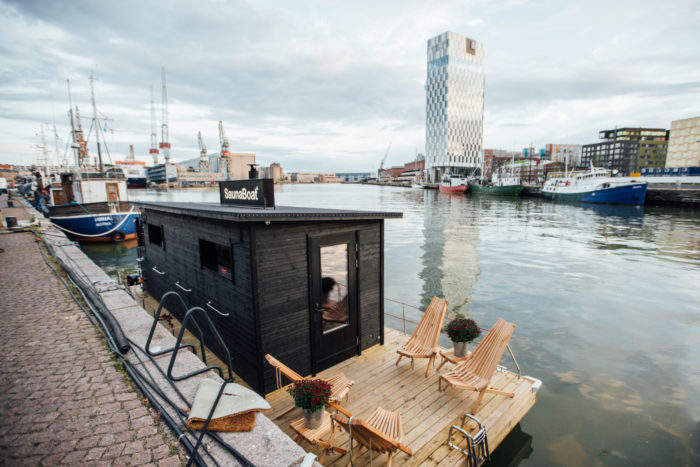 A raft with a small wooden sauna building and sundeck is docked in a Helsinki harbour.