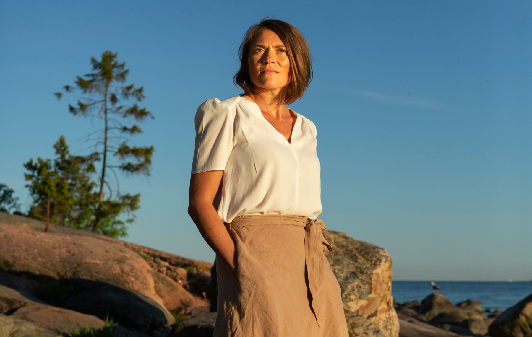 A woman stands in front of a rocky seacoast.